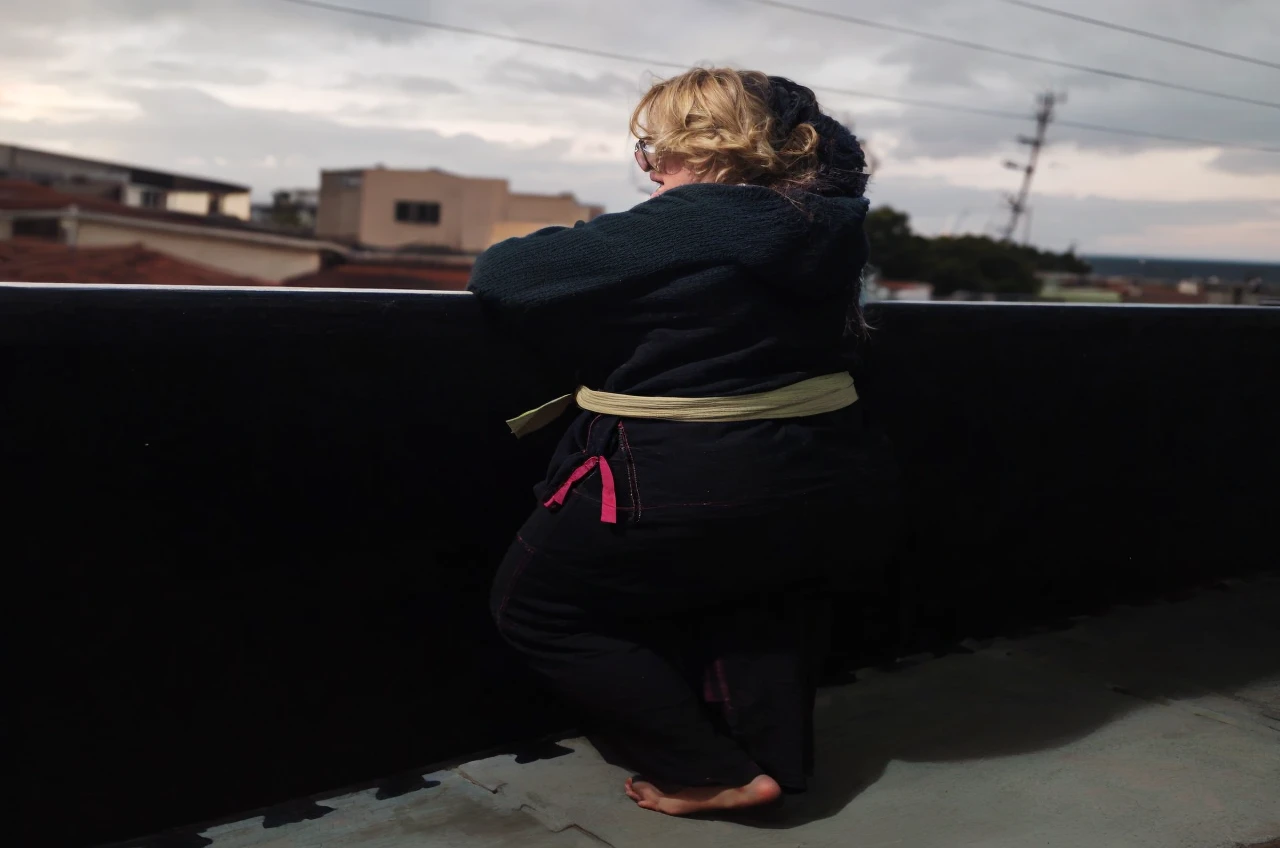 Slightly chubby blonde woman, of square glasses, blondie hair, clear green eyes, on the balcony looking at the sea to the horizon at sunset