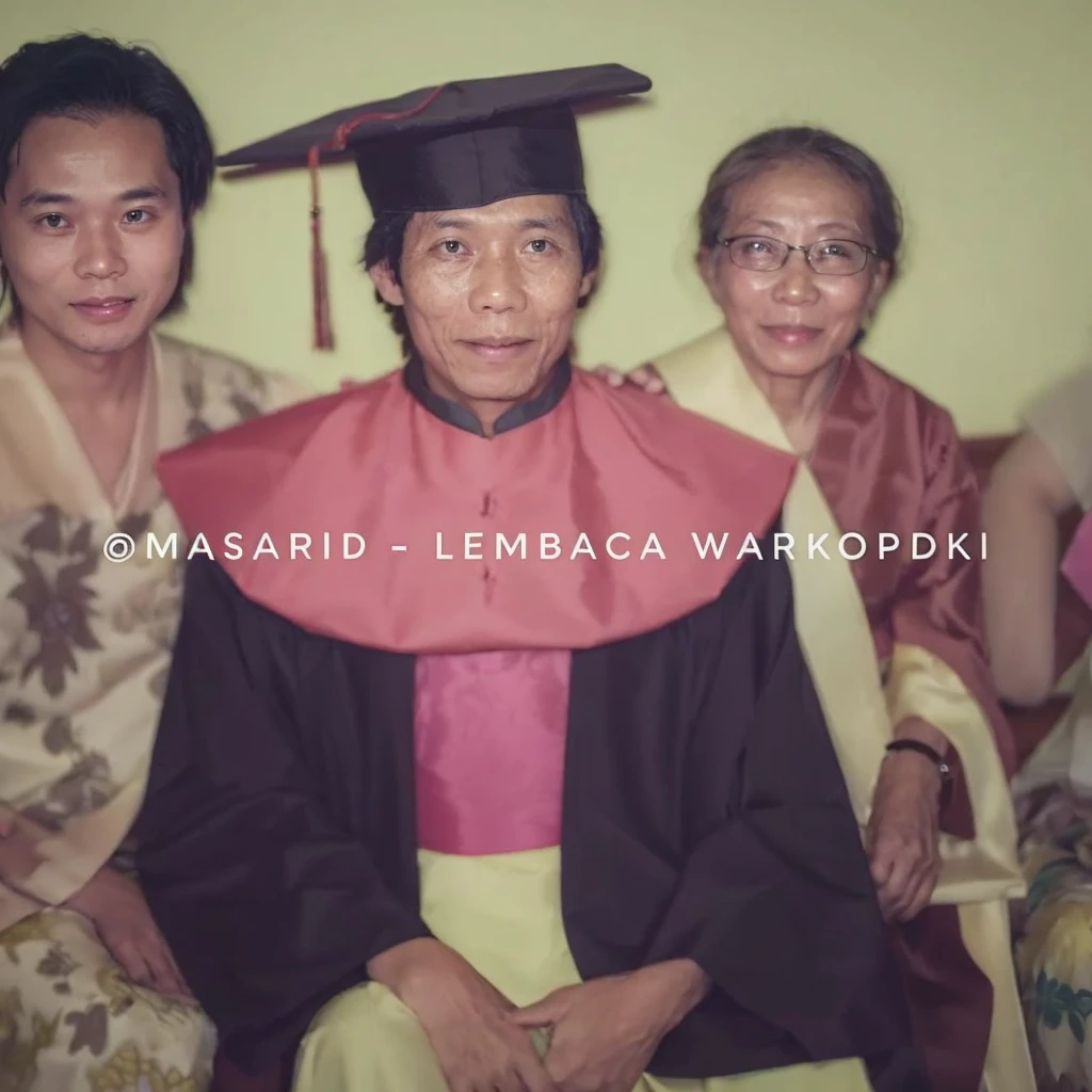 The image captures a heartwarming moment of a family celebrating a graduation. The graduate is seated between his mother and father, all three dressed in traditional Indonesian attire. The family proudly poses for a picture, with the graduate at the center of their joy.