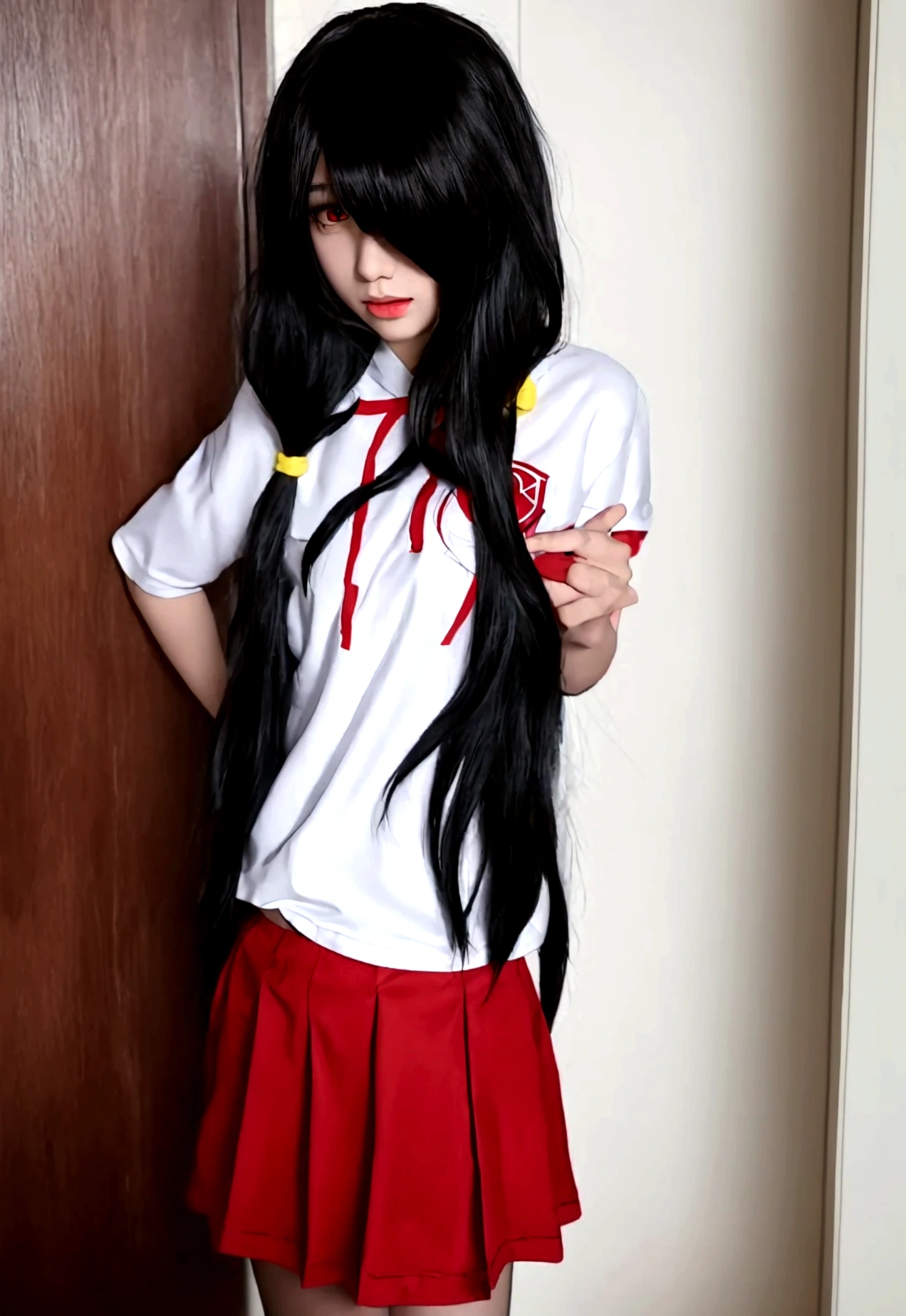 ultra-detailed,highly detailed,best quality,masterpiece,illustration, 
The image features an anime-style character with long, dark hair and heterochromatic eyes—one red and one yellow. The character is wearing a white sports shirt with red accents and a matching red skirt. The setting appears to be a locker room or a sports equipment room, as there are clothes and uniforms hanging in the background. The character's pose, with hands on hips and a slightly tilted head, suggests confidence or curiosity. The lighting is soft, casting gentle shadows and giving the scene a somewhat realistic touch despite its animated nature.