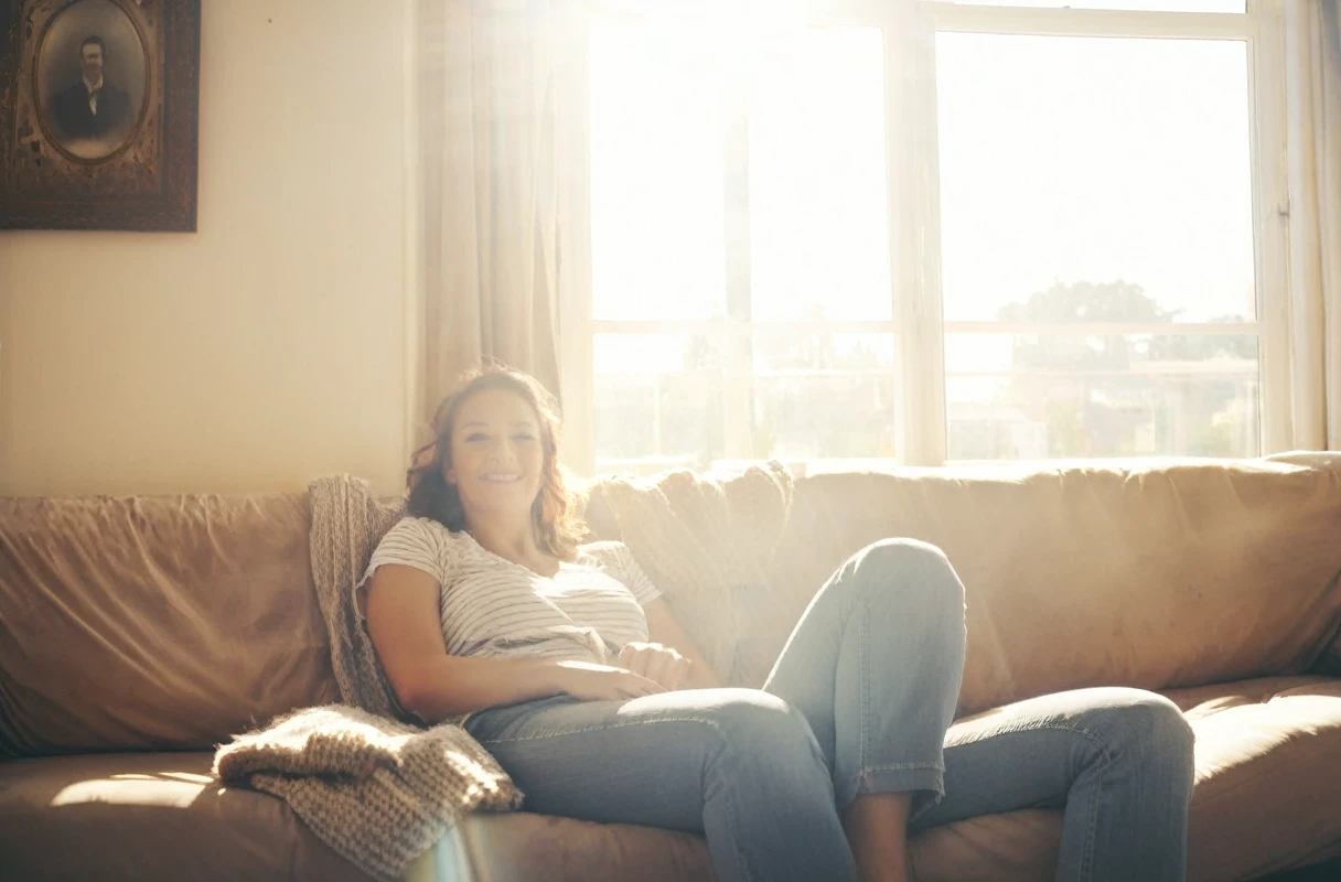 a photograph of highest quality a woman is sitting on her couch, photograph, 100mm, zoom, sunlight, indoors, happy, haze, atmosphere