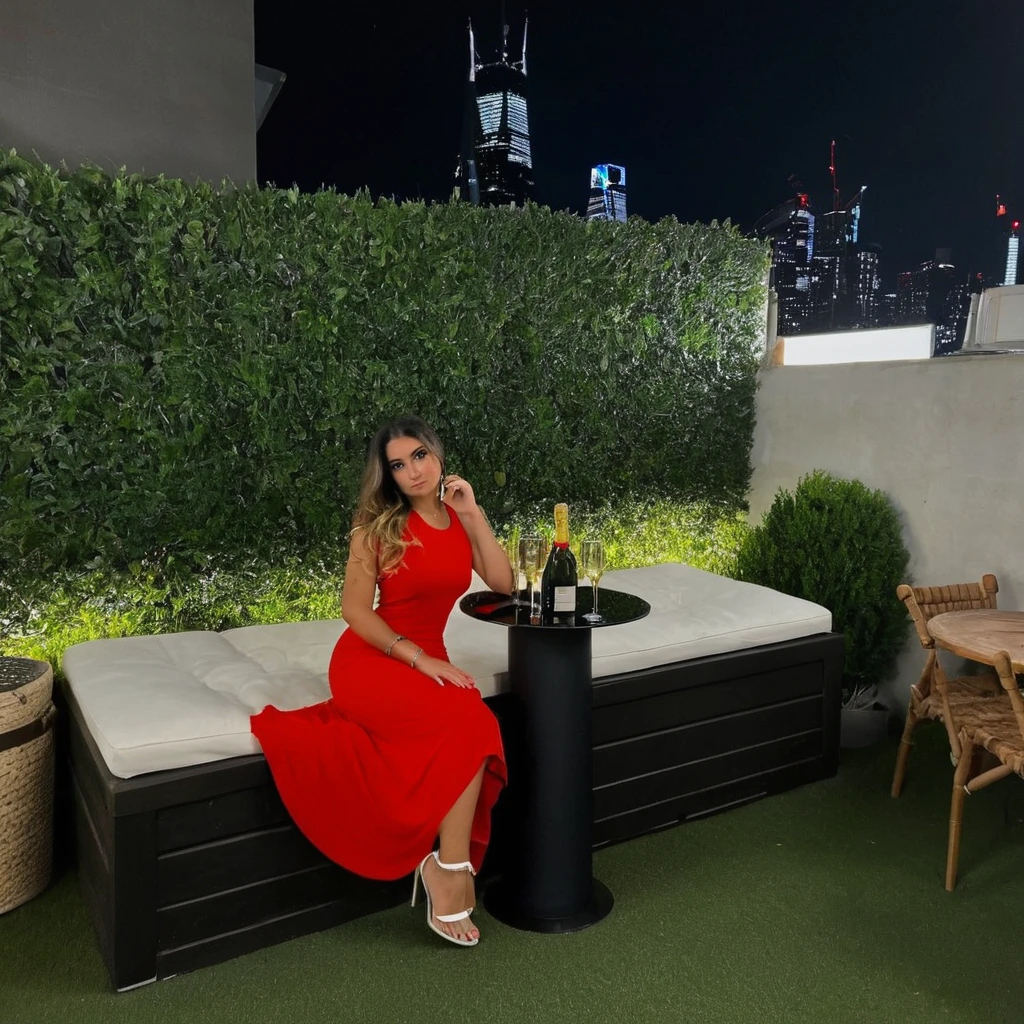 une femme incroyablement séduisante dans un cadre parfait pour un post Instagram. La femme a une silhouette élancée et porte une robe moulante élégante de couleur rouge vif avec des découpes audacieuses et des détails scintillants. Ses cheveux sont longs, lisses et noir corbeau, tombant avec grâce sur ses épaules. Elle porte un maquillage glamour avec un rouge à lèvres rouge profond et un eye-liner prononcé. La femme pose de manière confiante, une main sur la hanche et l'autre tenant une coupe de champagne, avec un sourire captivant et irrésistiblement séduisant sur son visage. Le décor est une terrasse ultra-moderne située sur le toit d'un gratte-ciel avec une vue panoramique sur la ville illuminée de nuit. La terrasse est équipée de meubles chics et luxueux, incluant des canapés en velours noir avec des coussins dorés et des tables basses en marbre blanc. Des lumières LED intégrées au sol et des lanternes design en verre fumé créent une ambiance intime et sophistiquée. À proximité, une table haute en marbre est ornée de bougies parfumées, de coupes de champagne pétillant et de plats gastronomiques, ajoutant une touche de luxe et de sensualité. Un jacuzzi à débordement illuminé par des LED ajoute une dimension de plaisir et d'élégance. Il y a également un bar à cocktails exclusif avec des barmans en smoking, prêts à servir des boissons raffinées garnies de fruits exotiques et de herbes aromatiques. La palette de couleurs est provocante et élégante, dominée par des tons de rouge, d'or et de noir, avec une ambiance générale de luxe, de séduction intense et de sophistication nocturne.