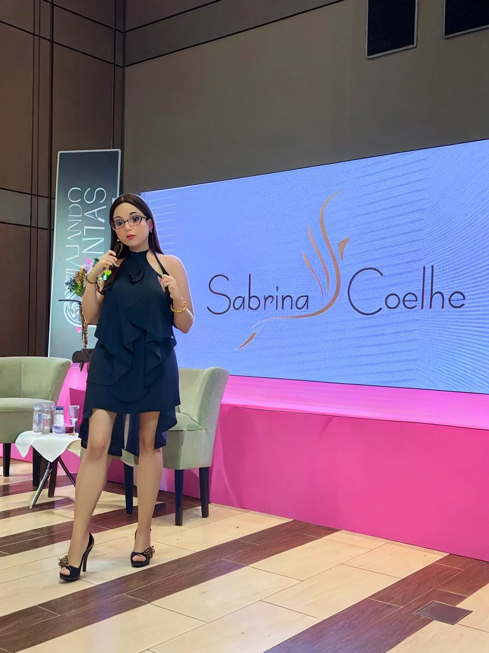 brunette woman in a black dress, long loose black hair, in prescription glasses, in high heels presenting a lecture, written on the screen sabrina coelho