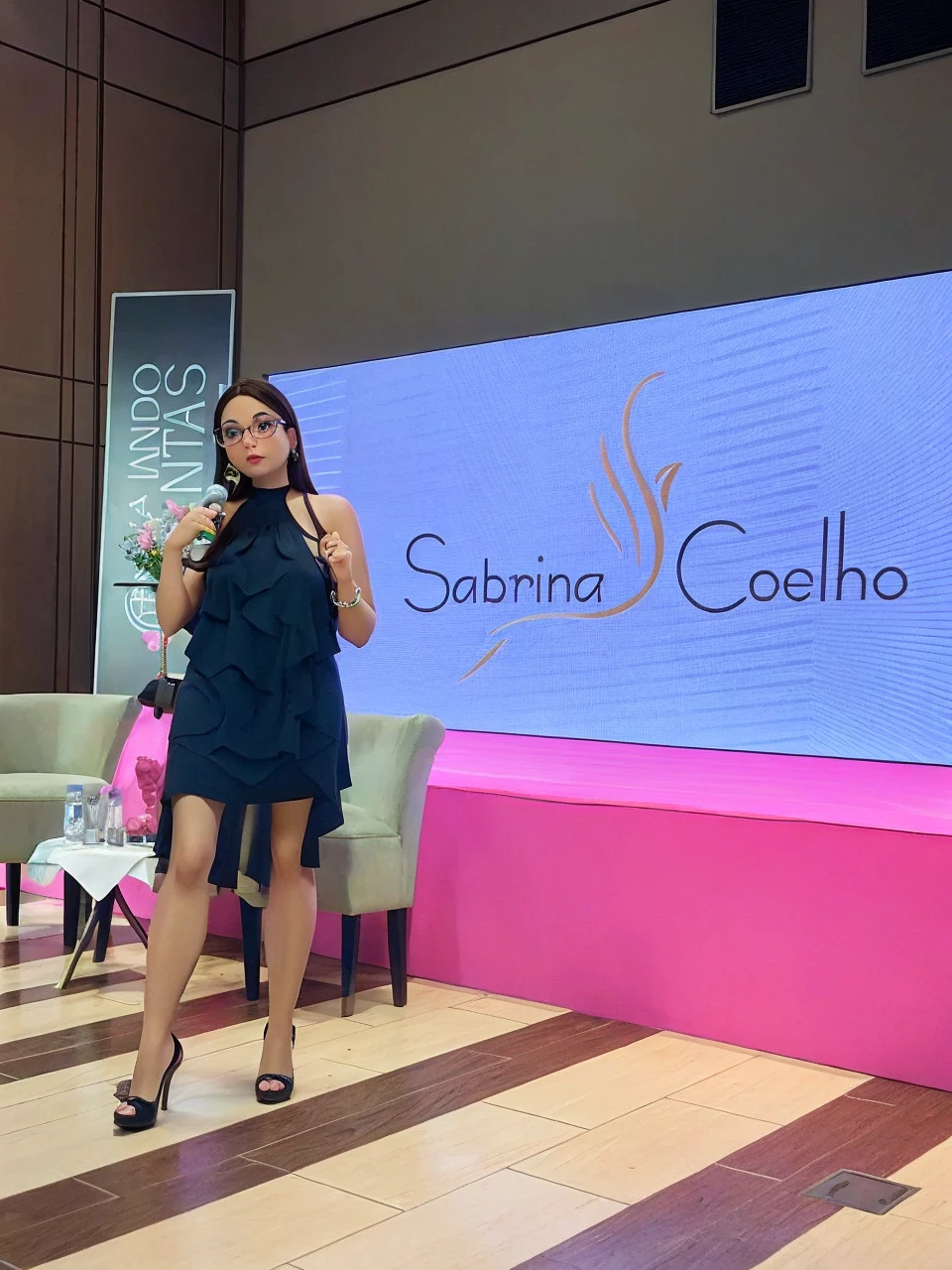 brunette woman in a black dress, long loose black hair, in prescription glasses, in high heels presenting a lecture, written on the screen sabrina coelho