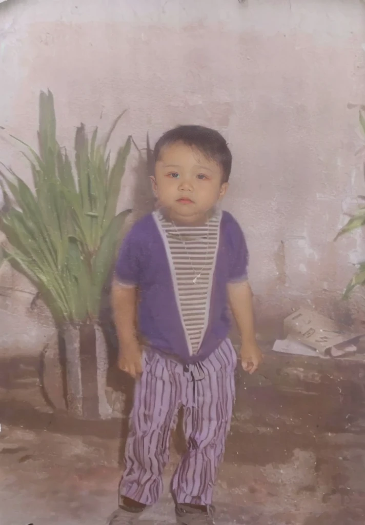 A boy wears a purple shirt with a striped pattern, striped pants, and black shoes, the wears a necklace, in the background there is a large pot with green plants and there is also a newspaper, a white wall.