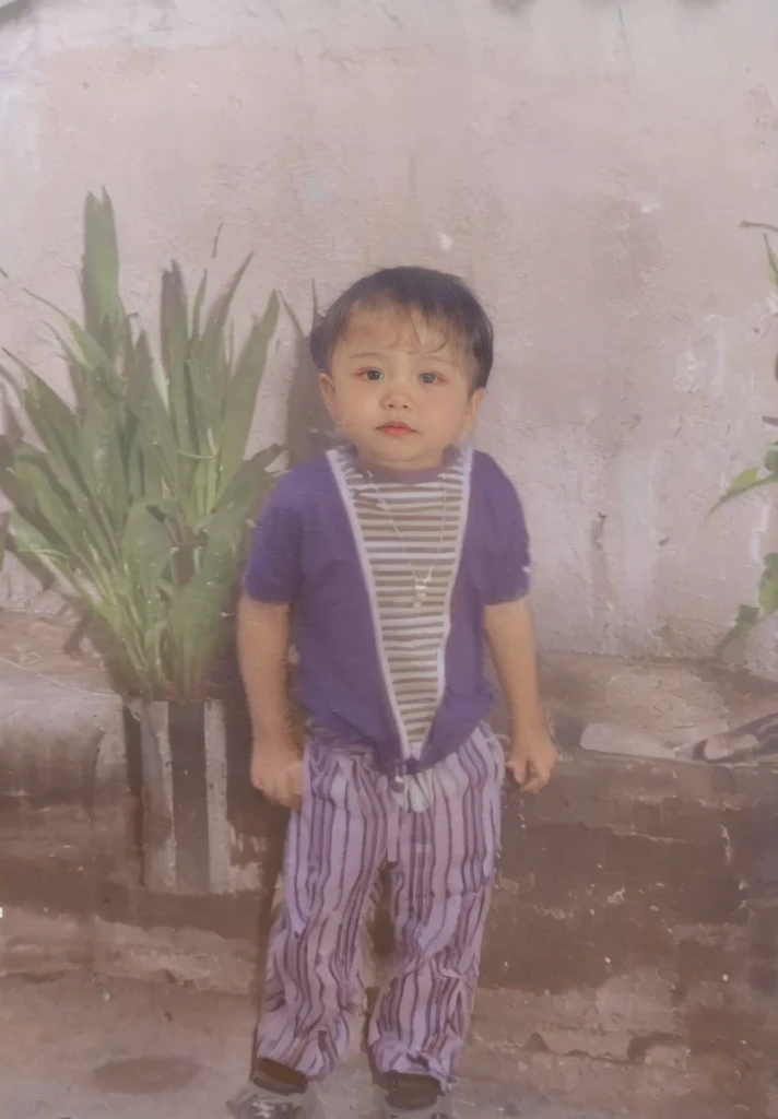 A boy wears a purple shirt with a striped pattern, striped pants, and black shoes, the  wears a necklace, in the background there is a large pot with green plants and there is also a newspaper, a white wall.