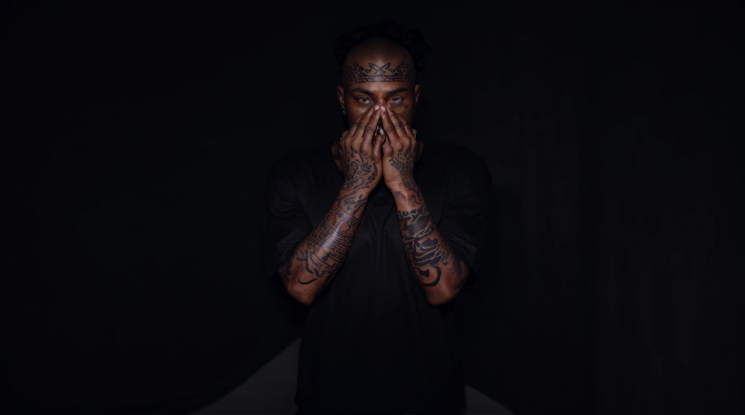 A dark-skinned man with tattoos on his arm covers his face except for his eyes, the eyes are grey, has a crown tattooed over his right eye, in Los Angeles at night, has a buzzcut and a black t-shirt