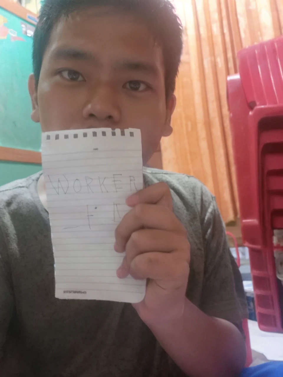 Indonesia guy holding a paper and the paper is facing front/in front of the perspective and the guy is holding the paper with 2 hands, in a messy room but not really messy, near perspective, the guy is in front of the camera