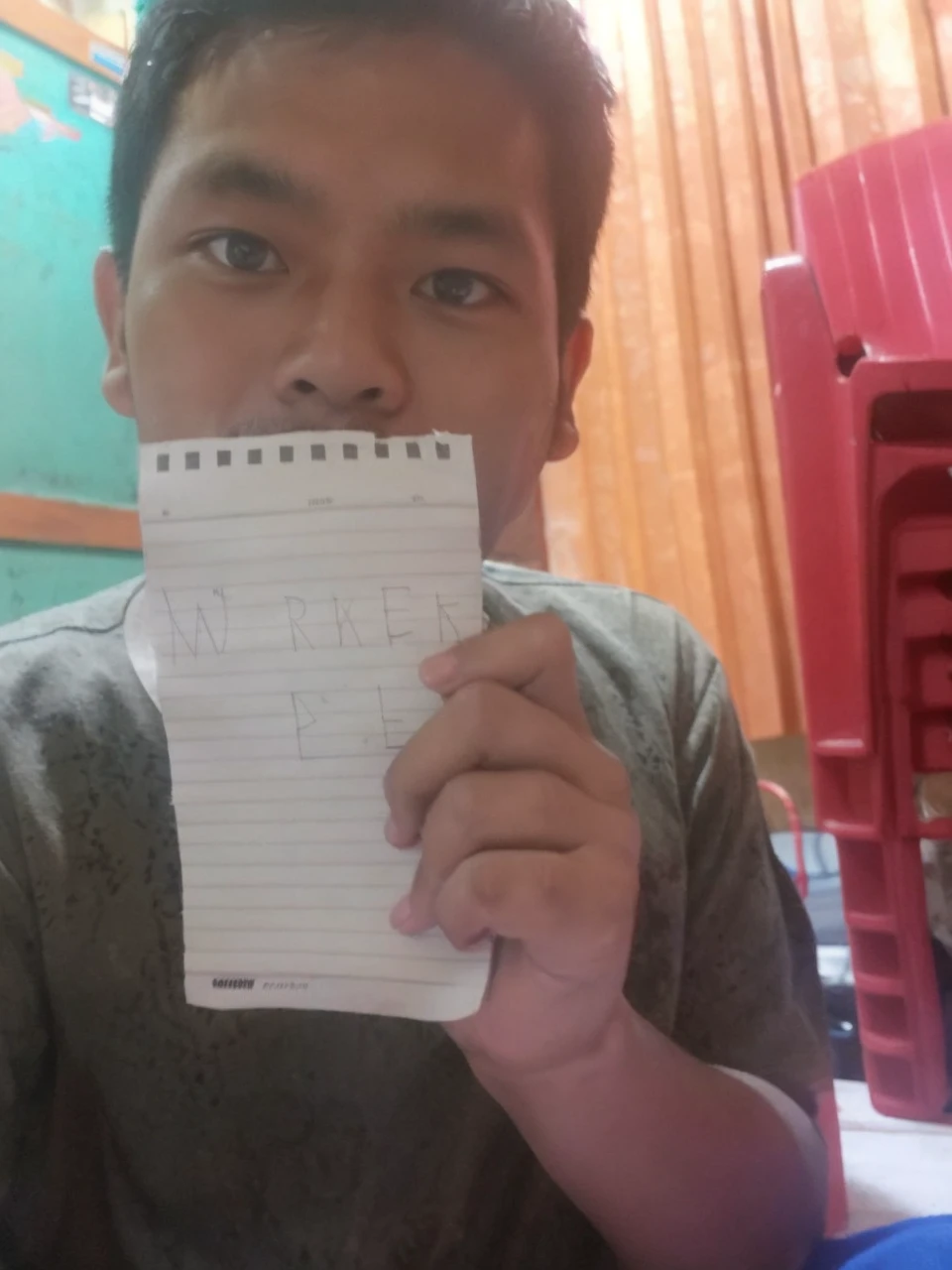 Indonesia guy holding a paper and the paper is facing front/in front of the perspective and the guy is holding the paper with 2 hands, in a messy room but not really messy, near perspective, the guy is in front of the camera
