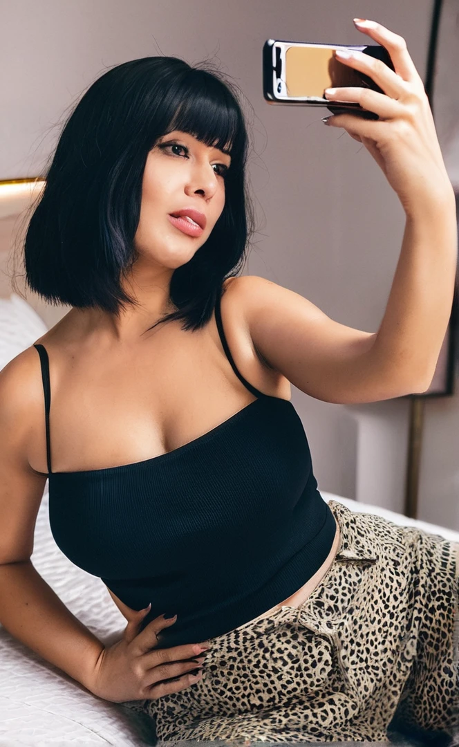 Woman taking a selfie in a bedroom with a bed and a black wardrobe., her navel is exposed, hands behind his body pose!, she has black hair with bangs, she has a round belly, fat and wavy, Thick, revealing outfit, hands behind his pose!, revealing outfit, 30 year old woman., lorena avarez