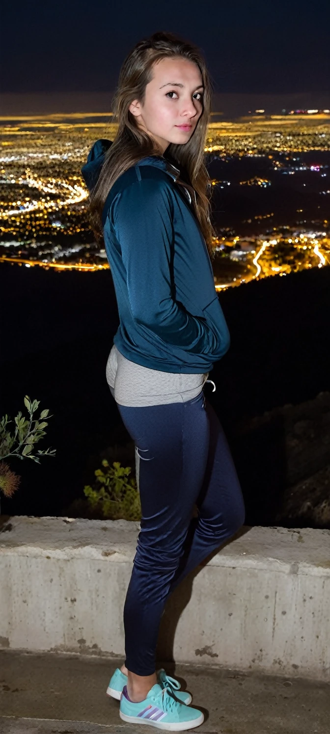 A girl with her hands in her pockets looking towards the me standing on the ledge of a mountain with a city in the background at night time
