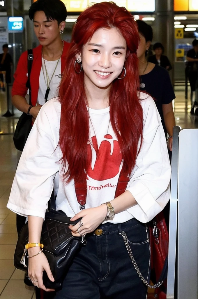 A Korean-Thai boy stands at the airport, smiles at the camera. He has red dyed hair and wears a hip-hop outfit. He also has an earring. Paparazzi take pictures of him. 