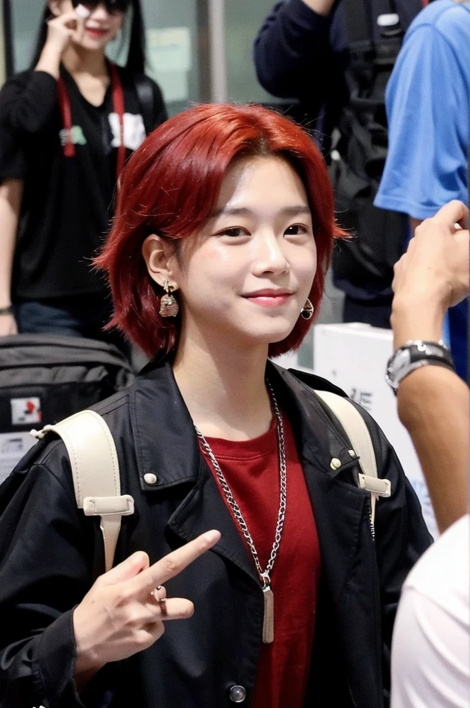 A Korean-Thai boy stands at the airport, smiles at the camera. He has red dyed hair and wears a hip-hop outfit. He also has an earring. Paparazzi take pictures of him. He checks Peace and has shoulder-length hair, which are wildly styled . 