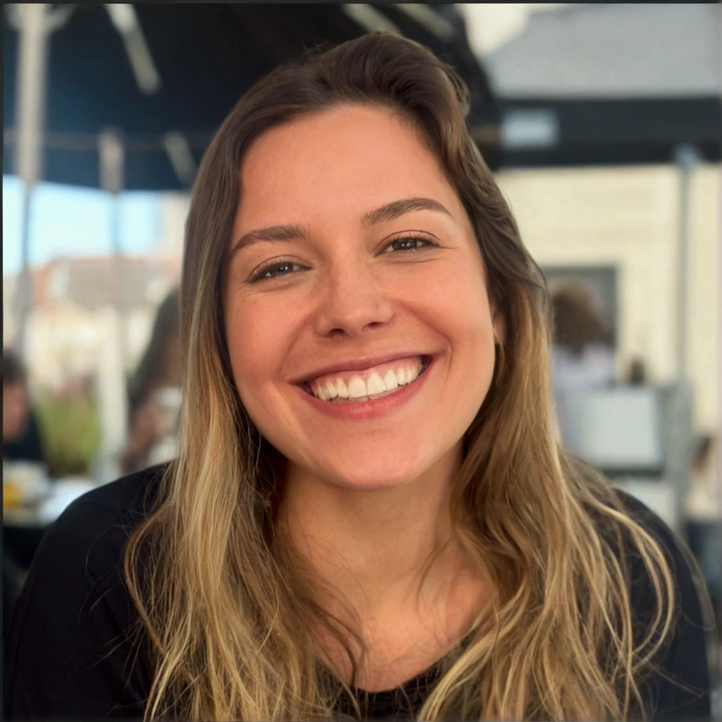 a woman with a wide, warm smile. She has long, blonde hair, that fall softly on your shoulders. She is wearing a black long-sleeved blouse. The woman is in an outdoor environment, probably in a cafe or restaurant, with a relaxed and illuminated atmosphere in the background. Her expression is one of joy and happiness, transmitting a feeling of well-being and positivity.