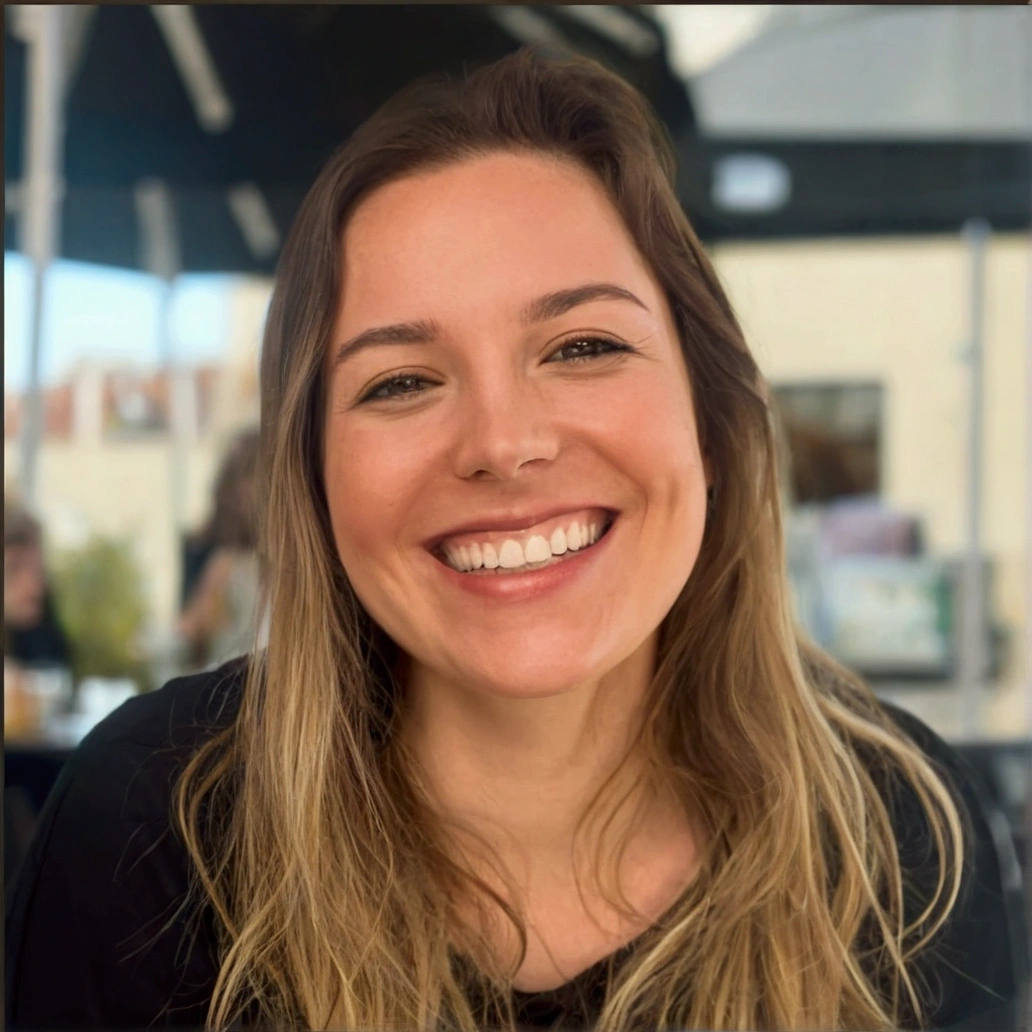 a woman with a wide, warm smile. She has long, blonde hair, that fall softly on your shoulders. She is wearing a black long-sleeved blouse. The woman is in an outdoor environment, probably in a cafe or restaurant, with a relaxed and illuminated atmosphere in the background. Her expression is one of joy and happiness, transmitting a feeling of well-being and positivity.