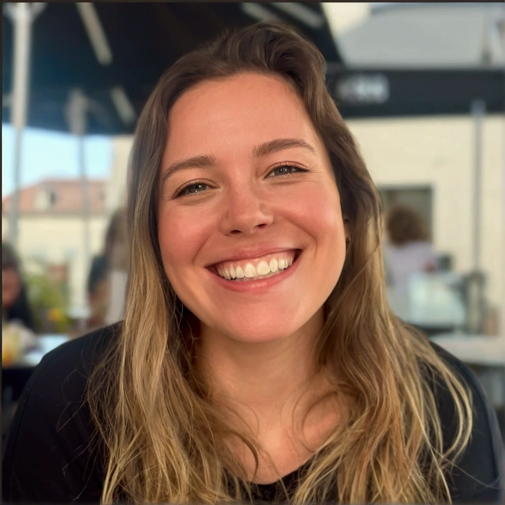 Cartoon style woman, pixar, a wide, warm smile. She has long, blonde hair, that fall softly on your shoulders. She is wearing a black long-sleeved blouse. The woman is in an outdoor environment, probably in a cafe or restaurant, with a relaxed and illuminated atmosphere in the background. Her expression is one of joy and happiness, transmitting a feeling of well-being and positivity.