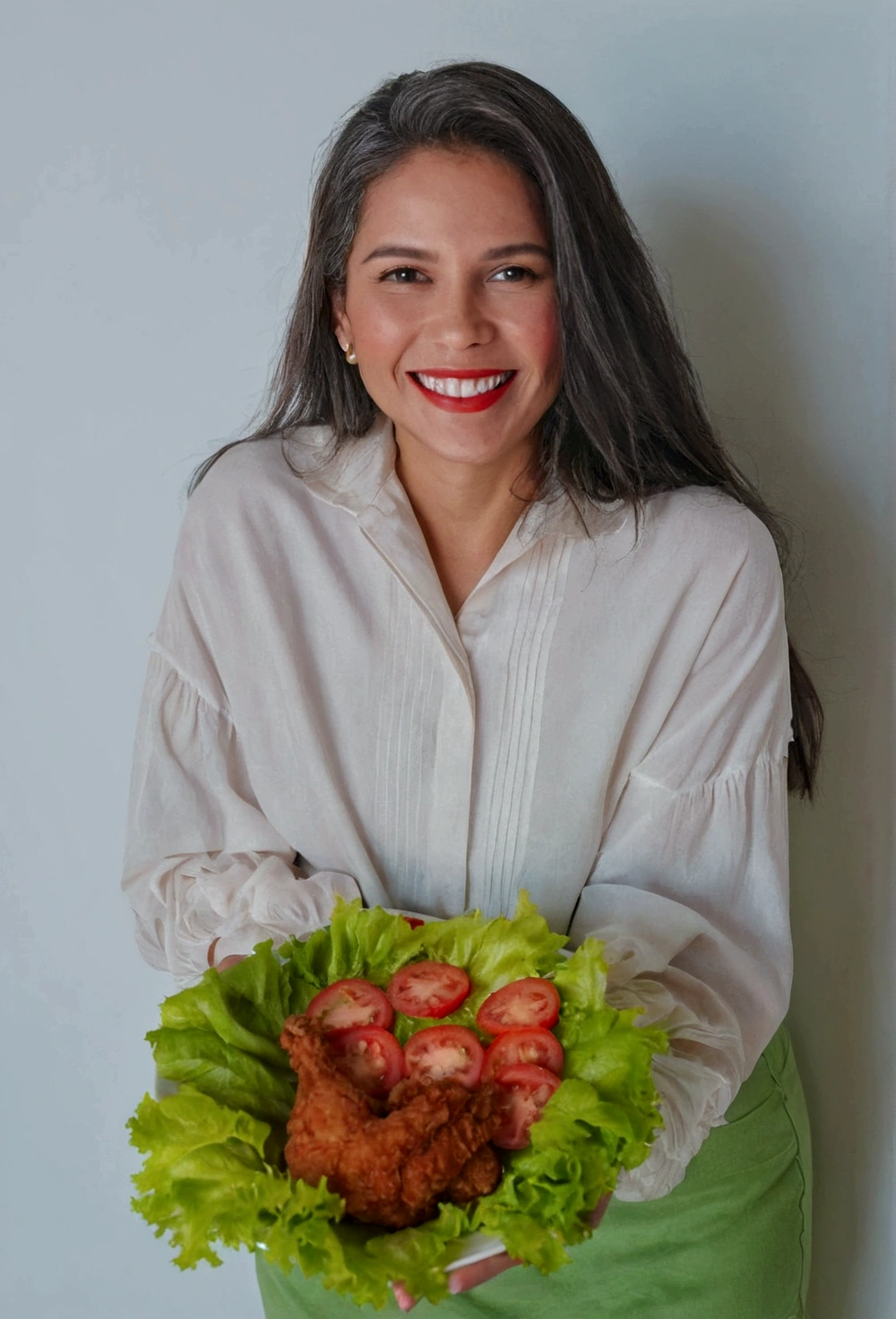 woman with medium wavy hair holding a plate of fried chicken with lettuce and cut tomatoes. She has a clear smile on her face. In the background there is a completely clear wall. Look at the model image and make it as close as possible to it, please.
