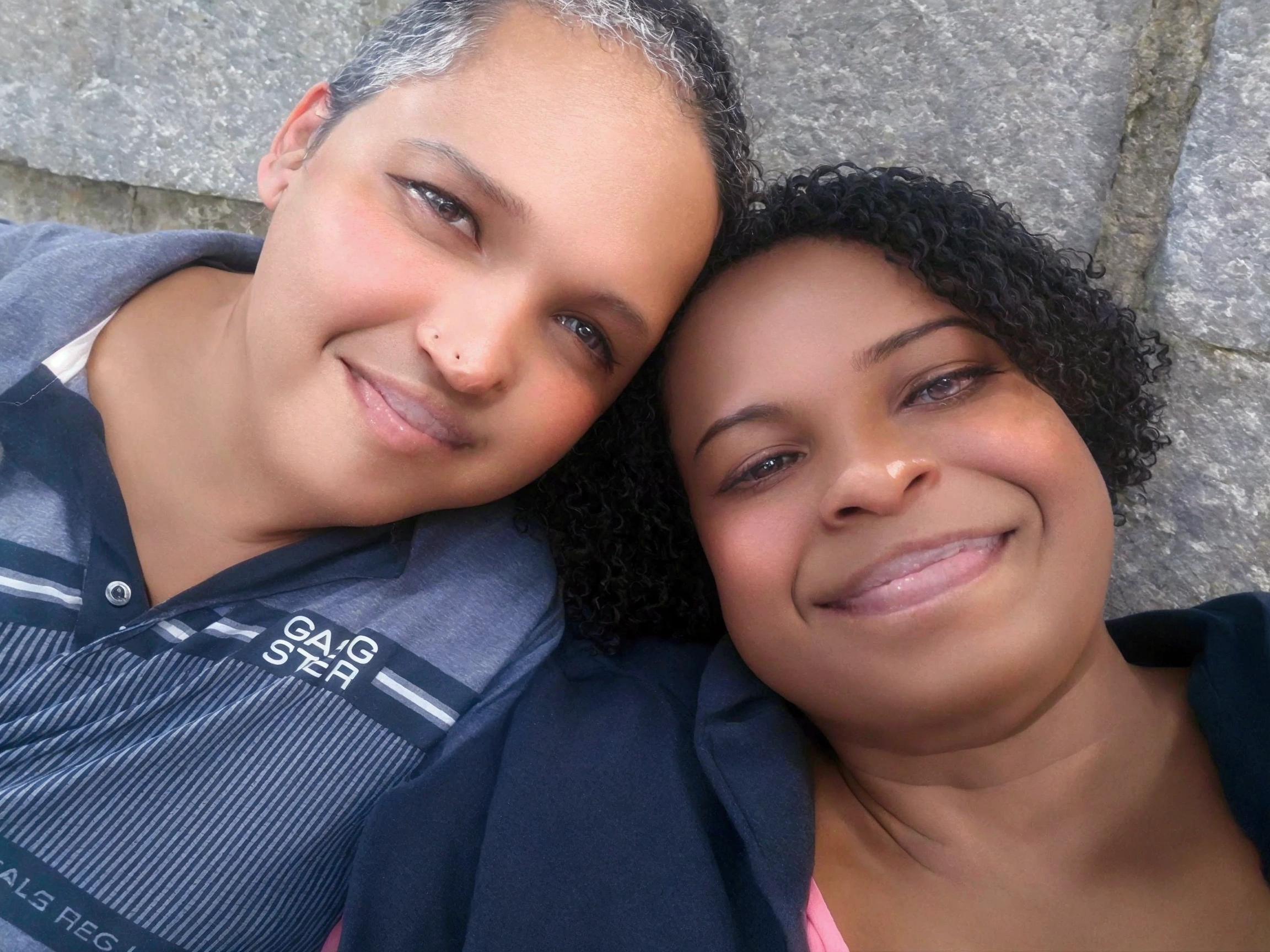 In this photo, there are two people smiling, one next to the other, appearing to be close and happy. The person on the left has light skin, short gray hair, and is wearing a dark gray polo shirt with white and gray horizontal stripes and the word "GAIGSTER" written on it. The person on the right has darker skin, short curly hair, and is wearing a dark blouse with a pink t-shirt underneath. The background of the image is made up of a stone wall. The atmosphere of the photo is one of closeness and joy.