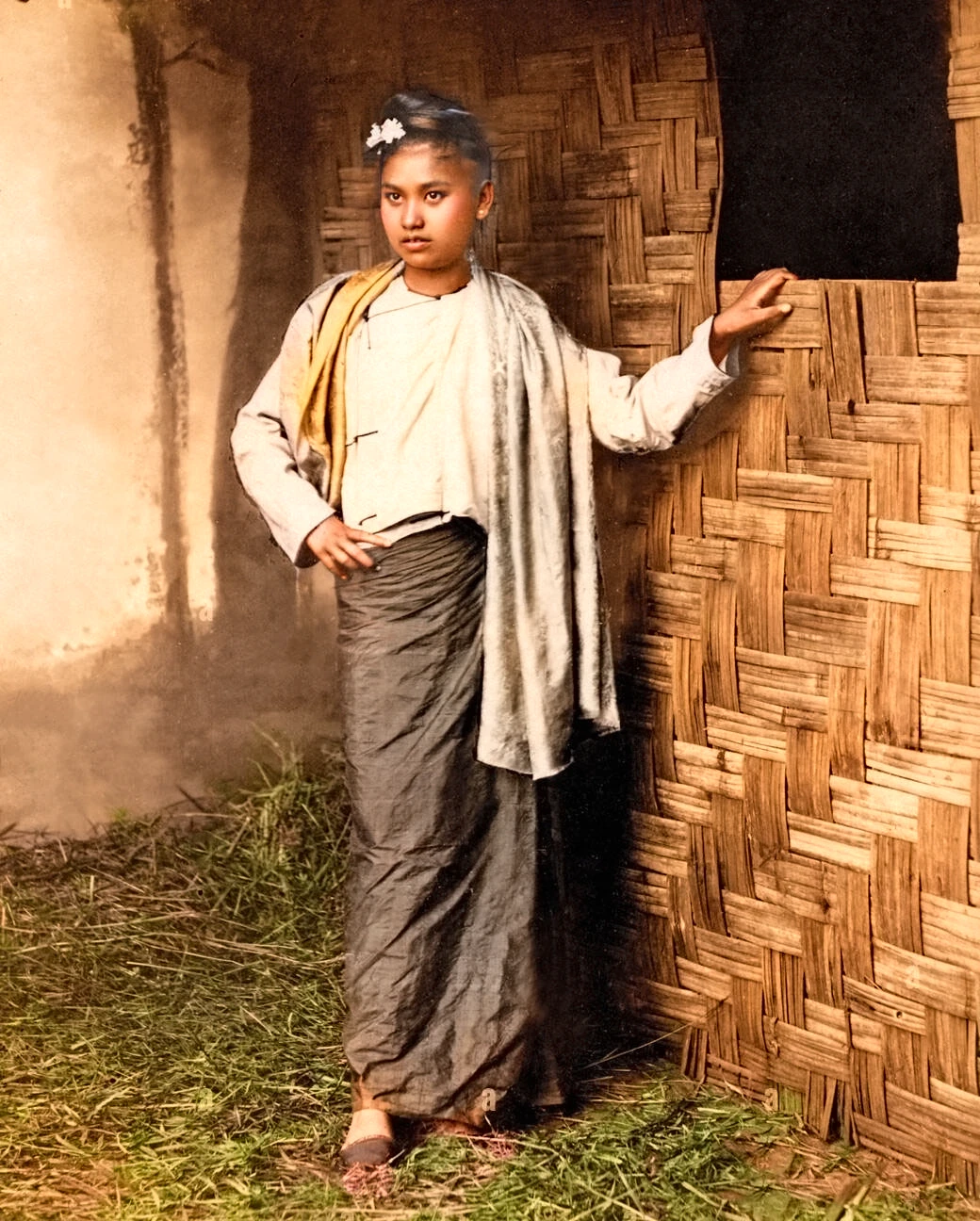 burmese woman standing in front of a bamboo rattan wall with a scarf. black hair. smooth skin. standing on dry grass.