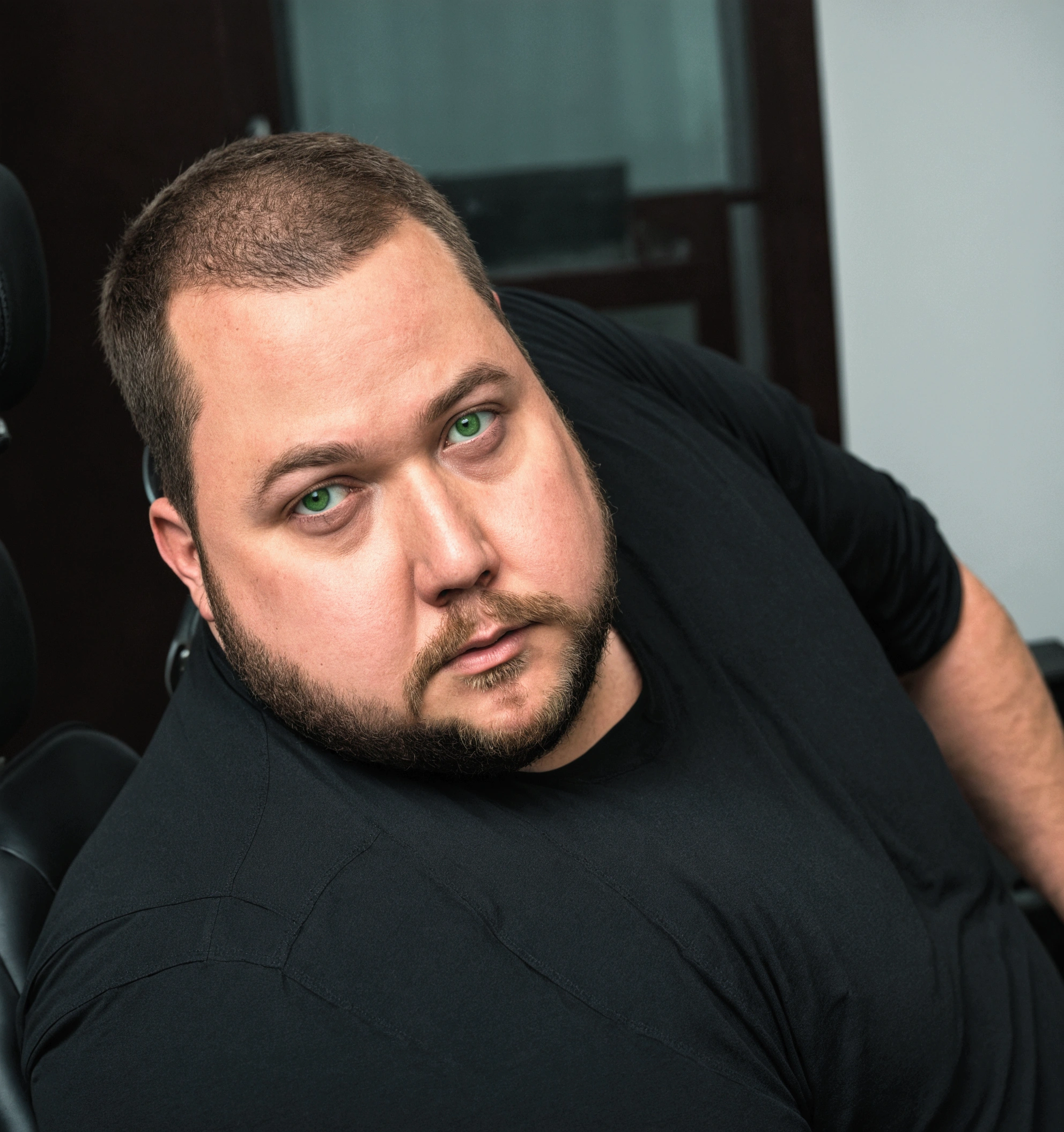 there is a man sitting in an office chair, chubby man with green eyes and brown, in black shirt, with beard, shaven hair