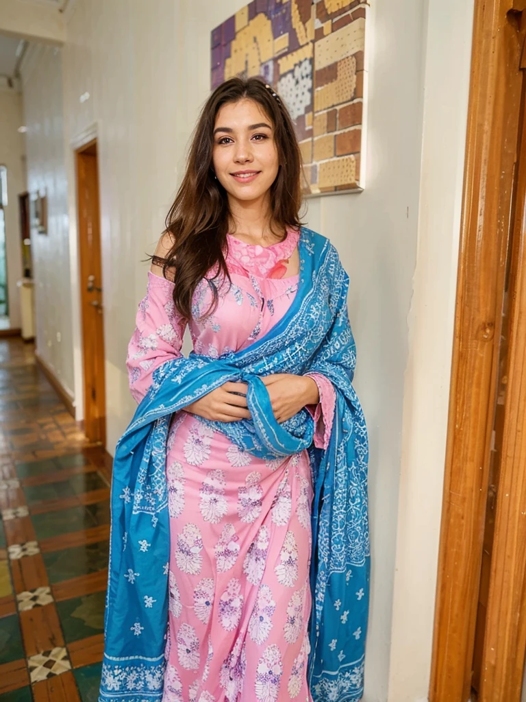 A woman in a traditional outfit stands indoors, smiling. She is wearing a pink dress adorned with white and purple patterns and has a blue patterned shawl draped over her shoulders. The background features a hallway with tiled flooring and a wall-mounted painting.