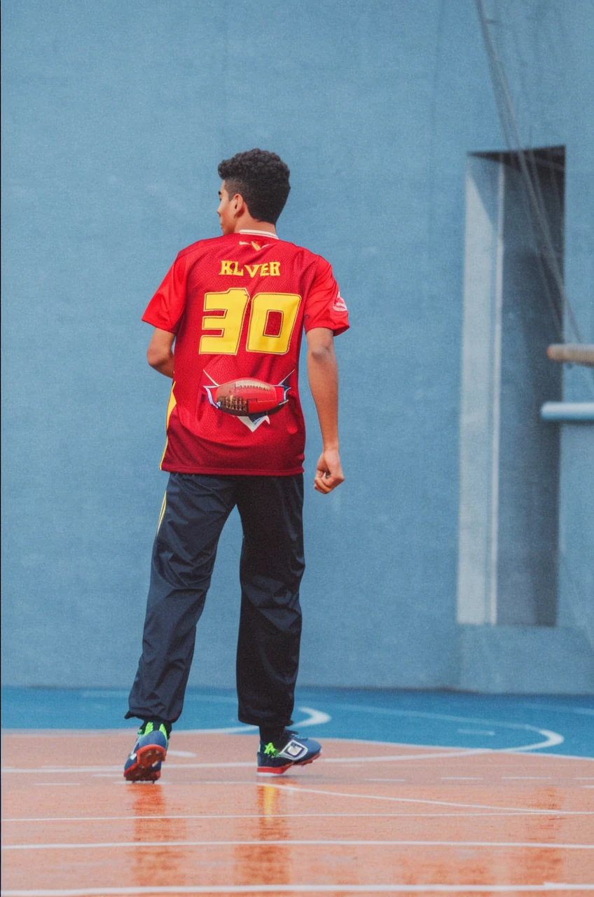 A young man with wavy hair, wearing a football shirt and shorts, is on a well-kept football field. He's in the middle of a move, with an expression of determination on his face, while running after the ball. Ao fundo, you can see the goalposts and some spectators in the stands, applauding and cheering. The sky is clear and blue, creating an atmosphere of enthusiasm and energy."