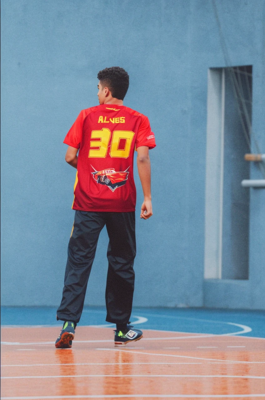 A young man with wavy hair, wearing a football shirt and shorts, is on a well-kept football field. He's in the middle of a move, with an expression of determination on his face, while running after the ball. Ao fundo, you can see the goalposts and some spectators in the stands, applauding and cheering. The sky is clear and blue, creating an atmosphere of enthusiasm and energy."