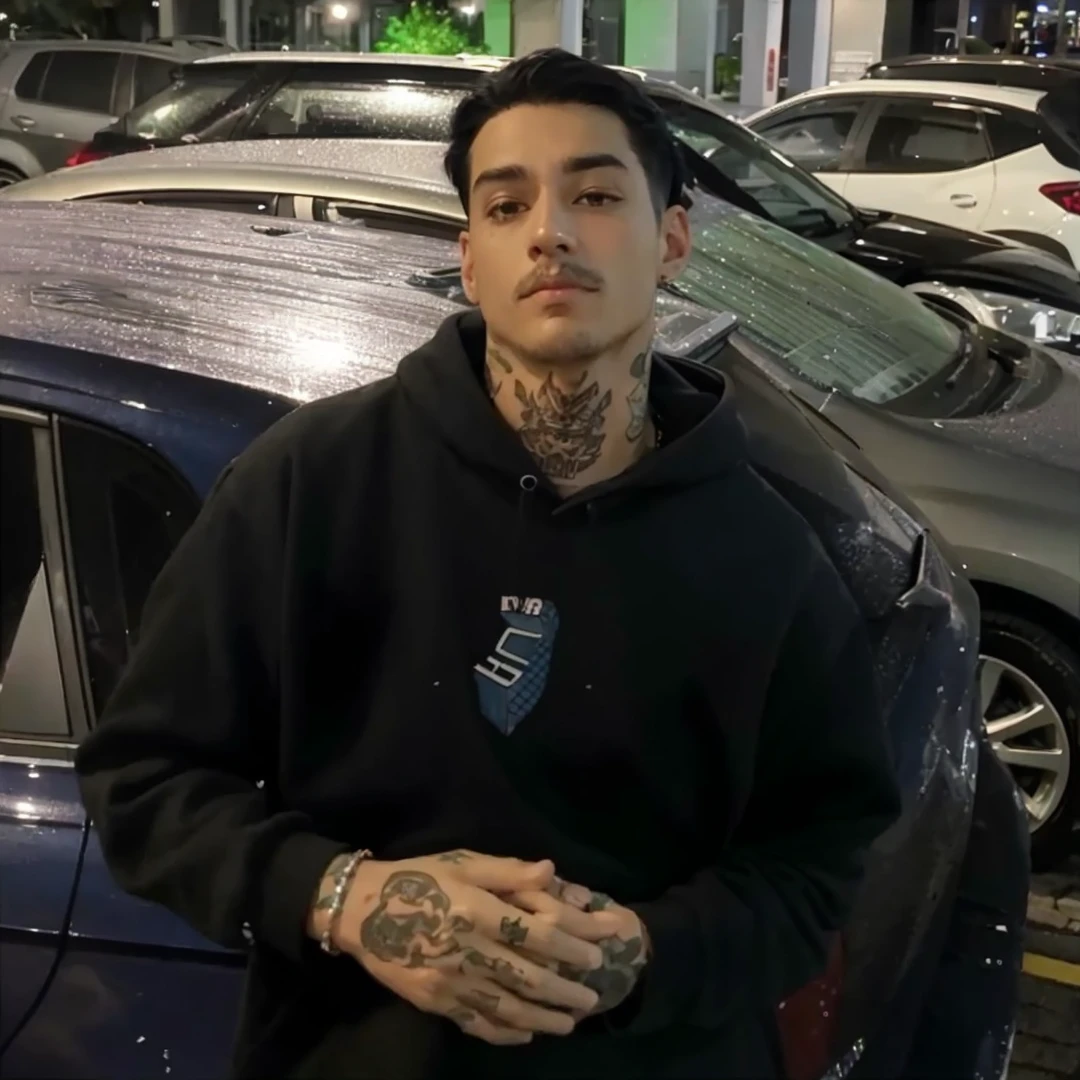 He has straight black hair, wears a black sweatshirt with a small blue print, there are cars behind him, he has tattoos and a thin mustache, appears to be 26 years old