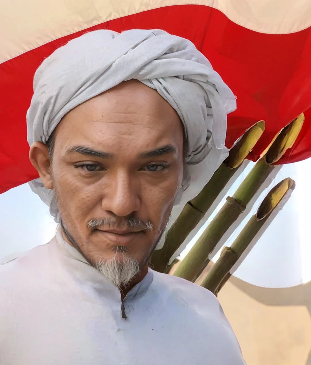 Indonesian Muslim man, 70 years old, wrinkled brown skin according to age. wearing a white patterned turban and Islamic clothes. clear black narrow eyes, thick gray eyebrows, thick mustache, long beard. big tall nose, big wide ears, oval face, naturally wrinkled forehead. mouth covered with thin lips. In the background of the Indonesian red and white flag, you can see three sharp bamboos on it. HD quality realistic images.
