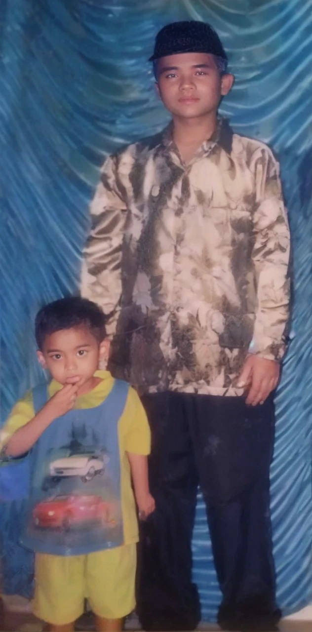 Boy in yellow blue clothes with a car image and yellow shorts, next to him a man wearing a black songkok with brown batik clothes with black pants, they are standing posing for the camera, the background is blue curtain