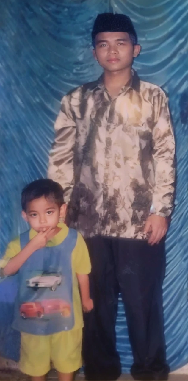 Boy in yellow blue clothes with a car image and yellow shorts, next to him a man wearing a black songkok with brown batik clothes with black pants, they are standing posing for the camera, the background is blue curtain