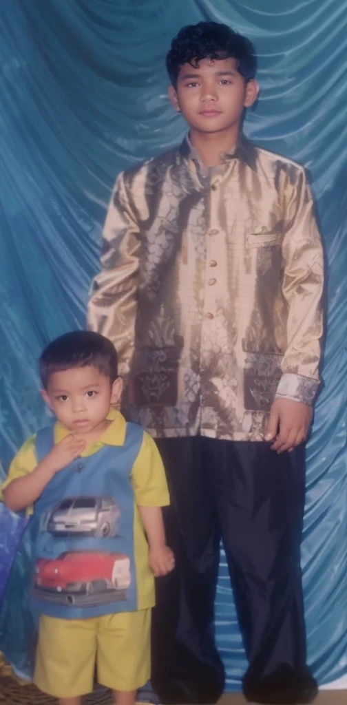 Boy in yellow blue clothes with a car image and yellow shorts, next to him a man wearing a black songkok with brown batik clothes with black pants, they are standing posing for the camera, the background is blue curtain