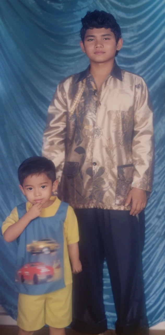 Boy in yellow blue clothes with a car image and yellow shorts, next to him a man wearing a black songkok with brown batik clothes with black pants, they are standing posing for the camera, the background is blue curtain