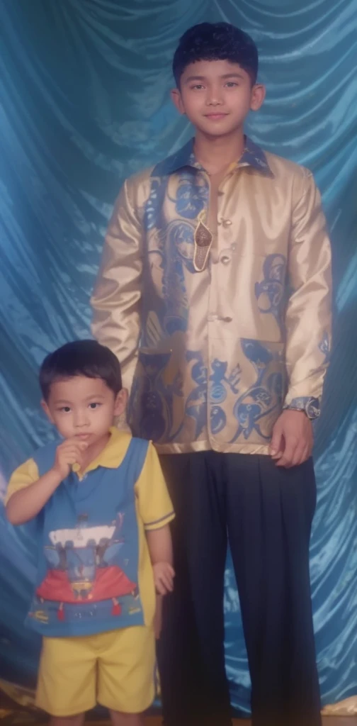 Boy in yellow blue clothes with a car image and yellow shorts, next to him a man wearing a black songkok with brown batik clothes with black pants, they are standing posing for the camera, the background is blue curtain