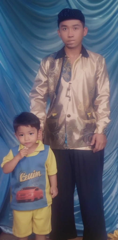 Boy in yellow blue clothes with a car image and yellow shorts, next to him a man wearing a black songkok with brown batik clothes with black pants, they are standing posing for the camera, the background is blue curtain