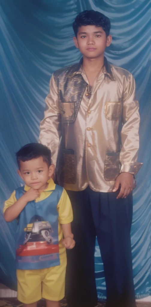 Boy in yellow blue clothes with a car image and yellow shorts, next to him a man wearing a black songkok with brown batik clothes with black pants, they are standing posing for the camera, the background is blue curtain