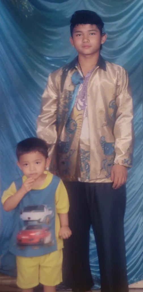 Boy in yellow blue clothes with a car image and yellow shorts, next to him a man wearing a black songkok with brown batik clothes with black pants, they are standing posing for the camera, the background is blue curtain