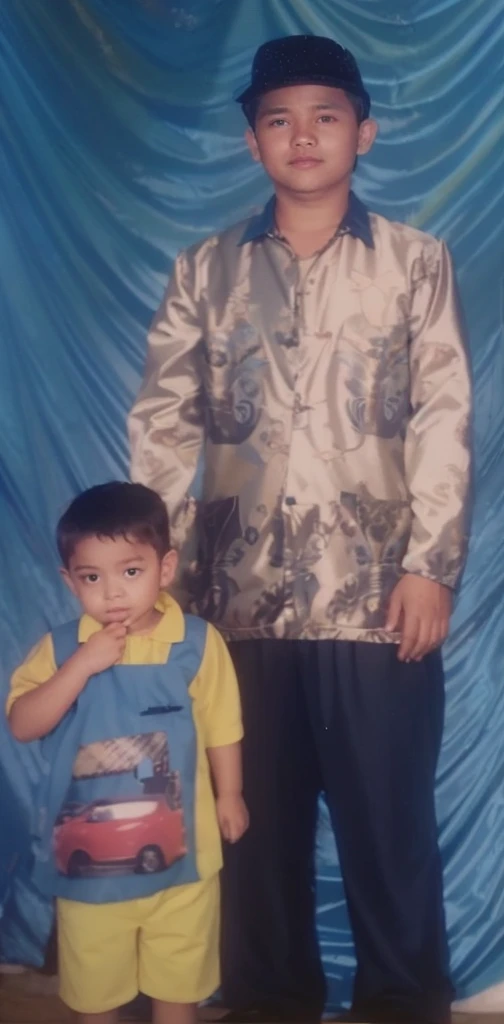 Boy in yellow blue clothes with a car image and yellow shorts, next to him a man wearing a black songkok with brown batik clothes with black pants, they are standing posing for the camera, the background is blue curtain