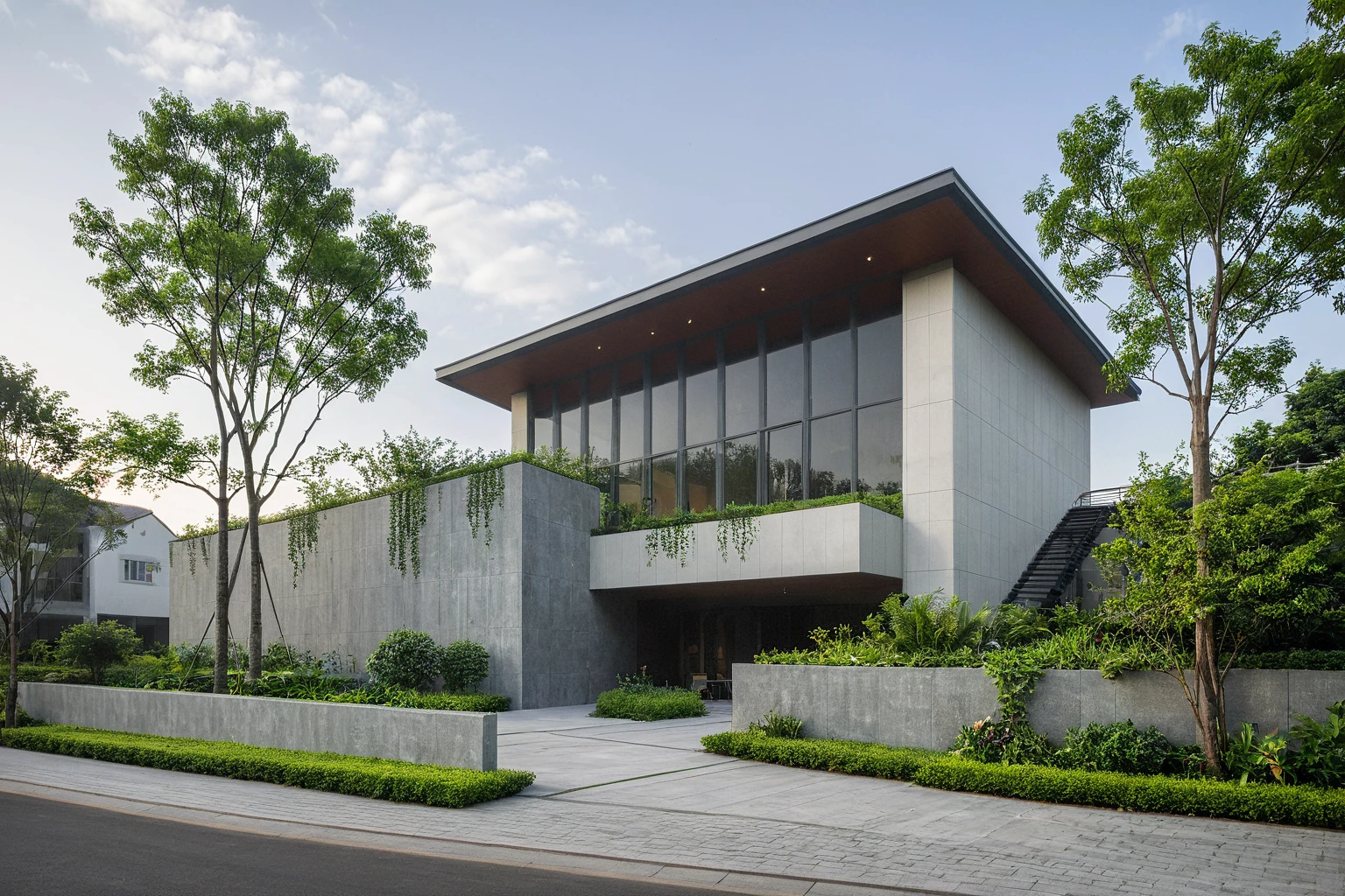 A modern villa situated on a street, basking in daylight. Surrounding the villa are lush tropical trees, green shrubs, and plants that add a vibrant touch of nature. The vivid colors of the vegetation create a striking contrast against the foggy and moody ambiance of the street. The villa boasts large glass windows and doors, constructed with high-quality, ultra-realistic glass that reflects the environment beautifully. The structure is made of solid concrete with a minimalist design, predominantly in dark grey tones. The interior glows with warm lighting, creating a welcoming atmosphere. The villa features modern materials and finishes of the best quality, resulting in a true masterpiece of contemporary architecture. The scene is enriched with streetscapes that blend seamlessly with the villa’s design, enhancing the overall aesthetic.
