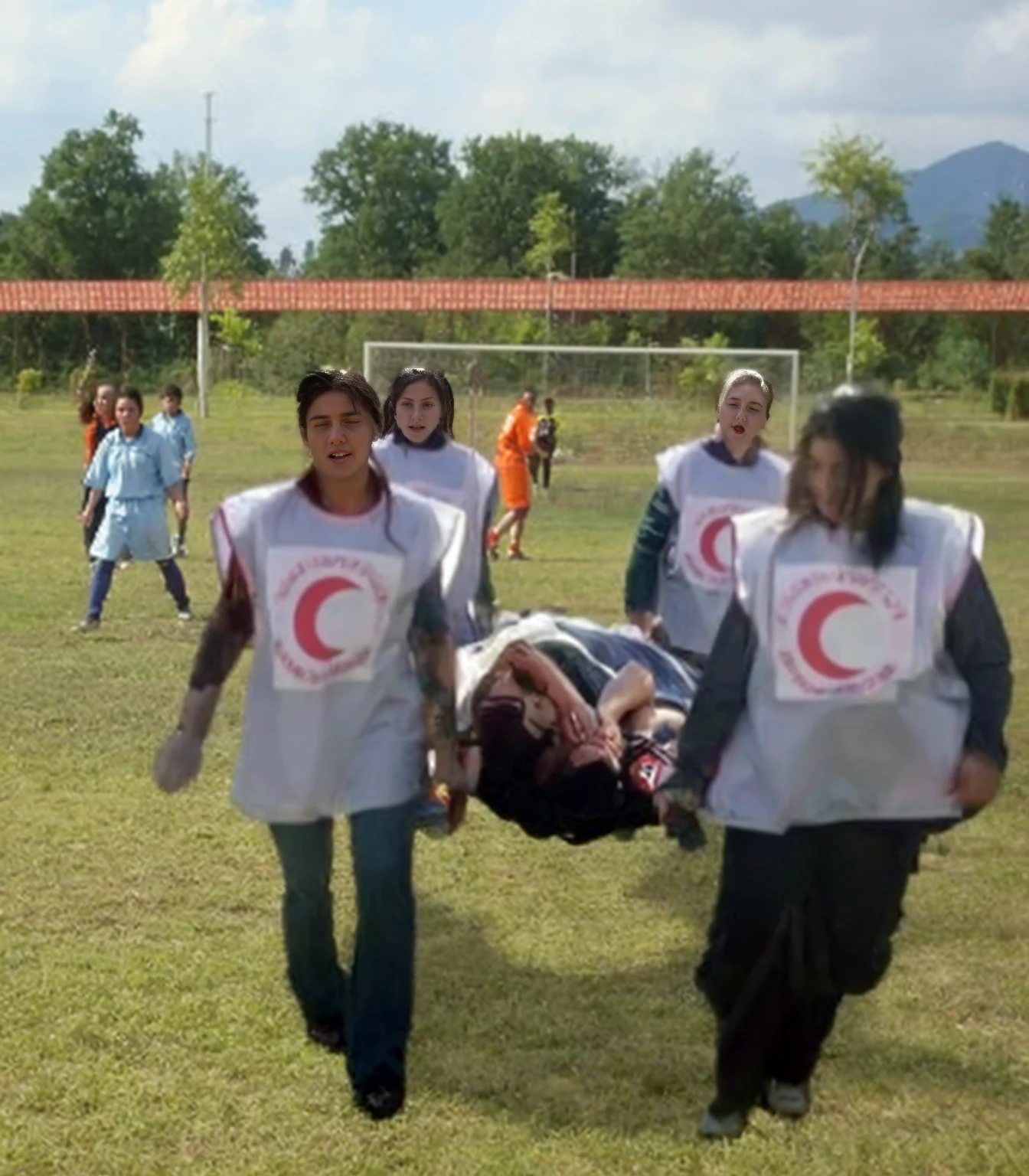 During a football match, an injured striker is carried off the pitch on a stretcher by four long-haired young women of the Red Crescent, Four long-haired young women in shiny Red Crescent uniforms carry a stretcher towards us on a football pitch, On the stretcher, an injured sporetler lies with his head facing us and has slapped his hands in front of his face in pain, An injured athlete lies on his back with his head first on a stretcher and has his hands on his face, Four long-haired young women in shiny uniforms carry the stretcher towards us, The young women all have very shocked expressions and scream and roar very loudly in horror
