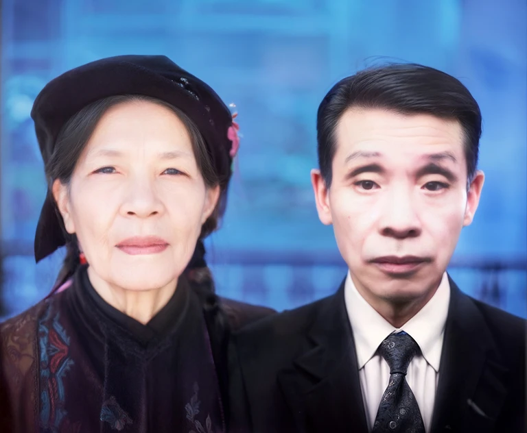 A portrait of an elderly Asian couple standing in front of the camera. The man is wearing a suit and tie, while the woman is dressed in traditional attire with a black headdress. Behind them is the garden of an old traditional Vietnamese house, adding a cultural and serene backdrop to the image.