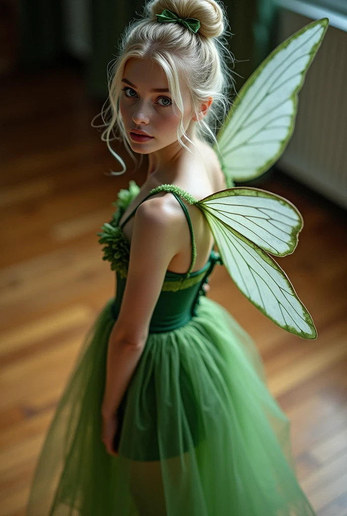 A shoclef looking 18 year old woman wearing a green fairy costume with white wings standing on a hard wood floor shot from above as she looks at the camera. Blonde hair in a bun