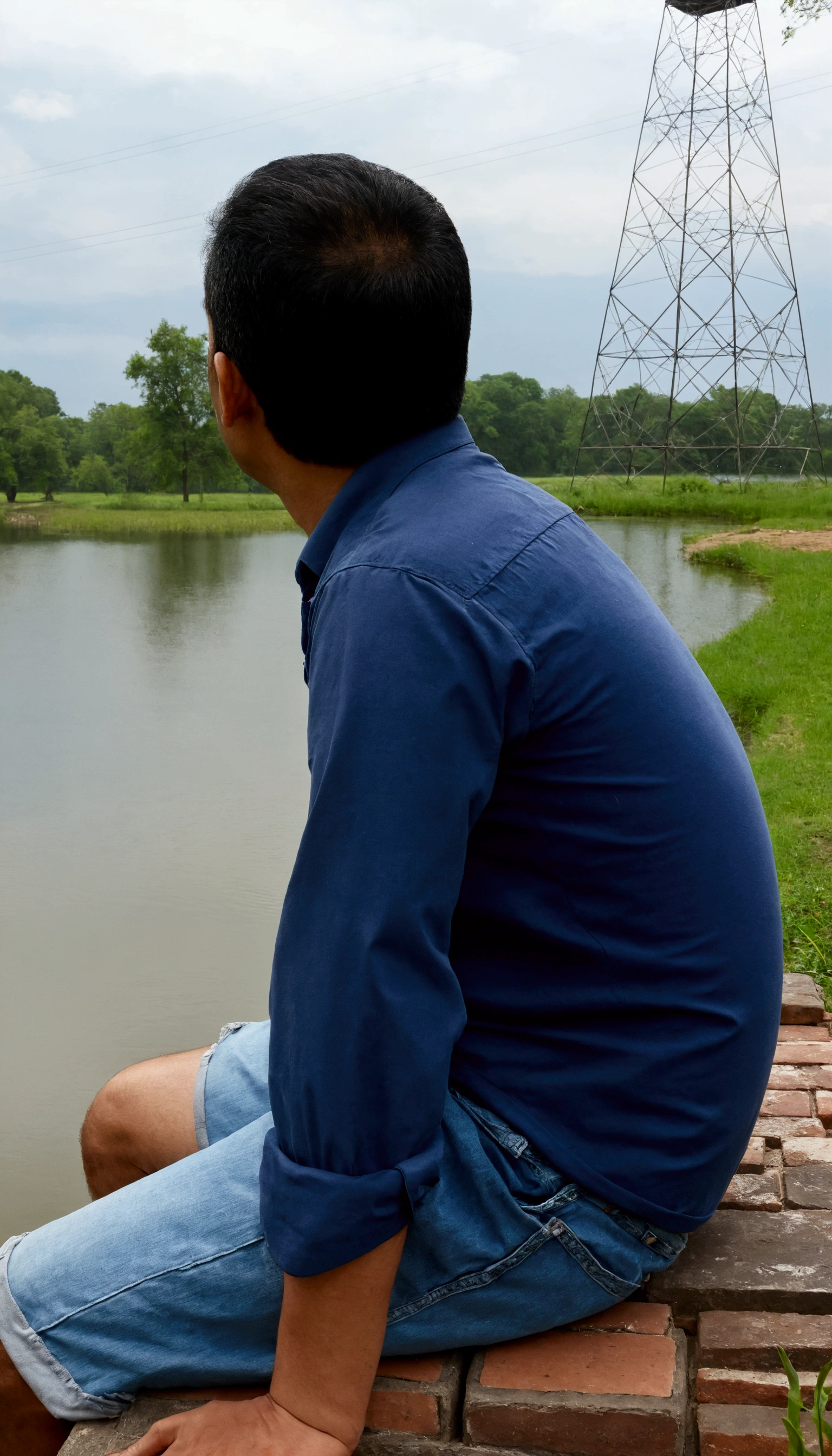 arafed man sitting on a brick wall looking out over a lake, sitting at a pond, sitting near a river, sitting in front of a lake, candid picture, lake in the background, river in front of him, river in the background, near a lake, distant full body view, candid photo, gazing at the water, standing in a pond, in lake
