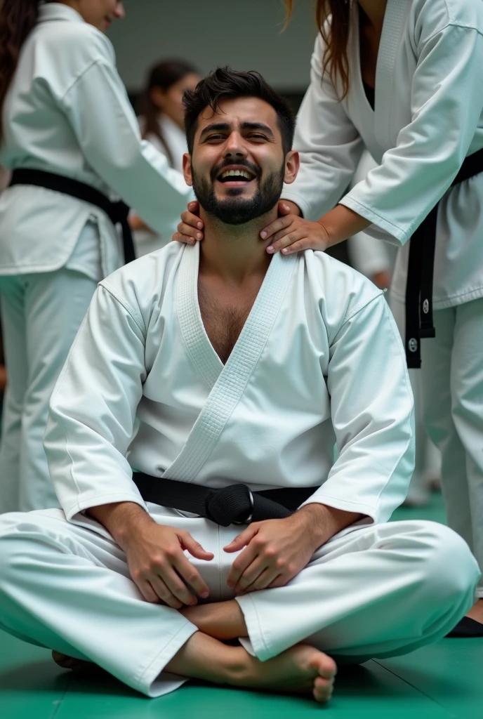 "A man wearing a taekwondo uniform is sitting on the floor performing a split, with a pained expression on his face. Another person is holding his waist. The floor is covered with a green mat, and there are other people in taekwondo uniforms around him."