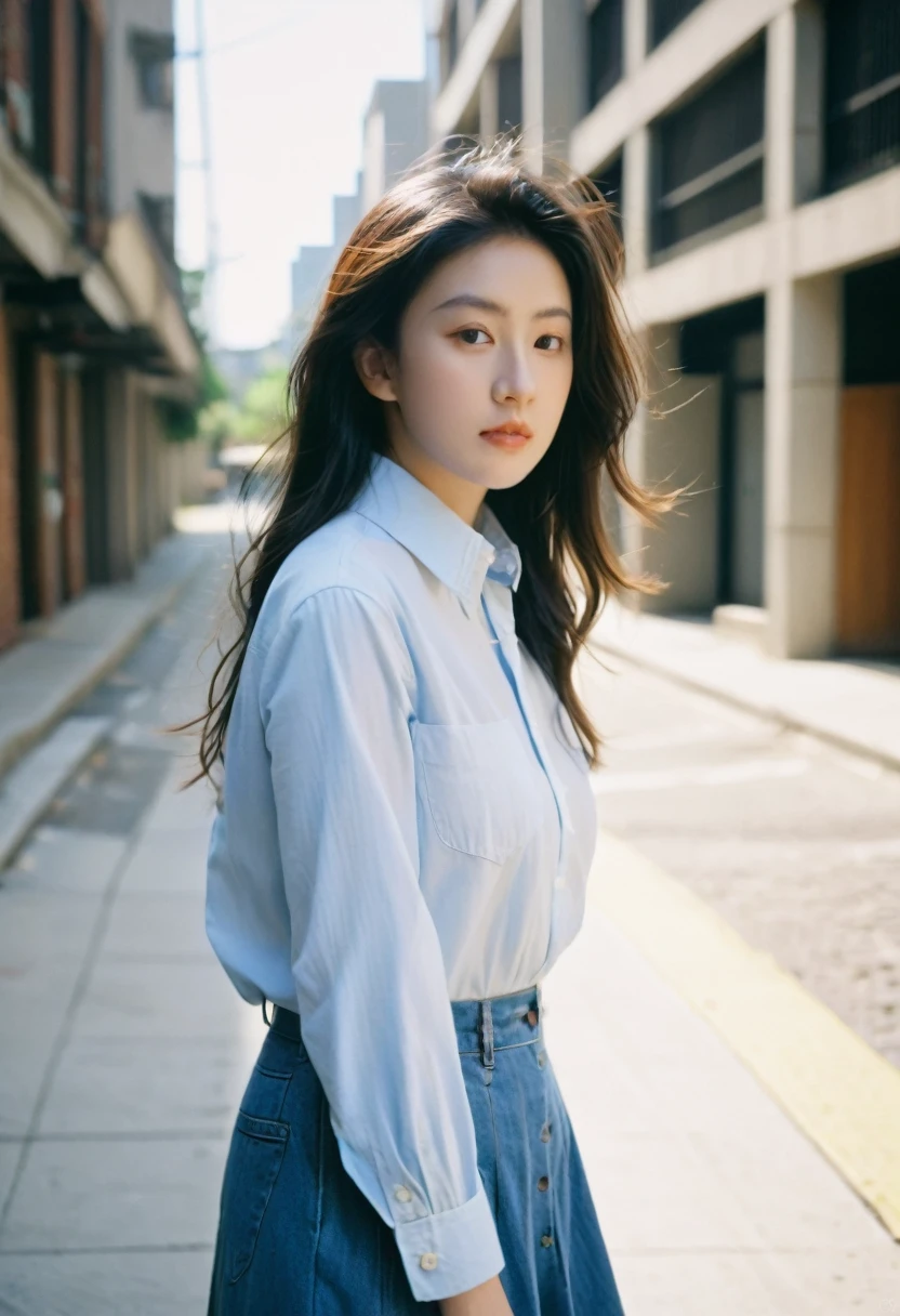 The image depicts a young woman with long, dark hair, looking directly at the camera. She is wearing a light-colored button-down shirt with a crest or emblem on the left chest, and a denim or light blue skirt. The photo is taken from a slightly elevated angle, giving a dynamic perspective. The background is blurred but appears to be an urban setting, possibly at night, with lights visible in the distance. The woman's expression is neutral or slightly serious, and her hands are positioned near her thighs as she slightly leans forward.