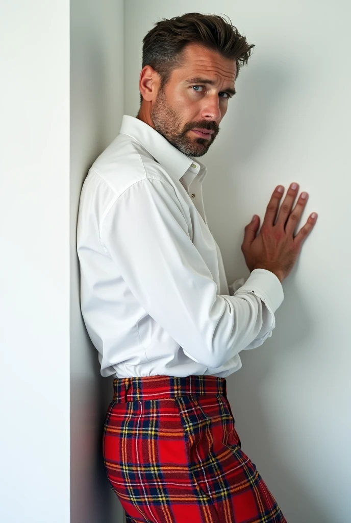 A white man, model type, at 33 years old, blue colored eyes, wearing a white shirt and a plaid skirt, standing with hands against the white wall of a room against the wall with her back to the camera and looking at the camera with a sexy look