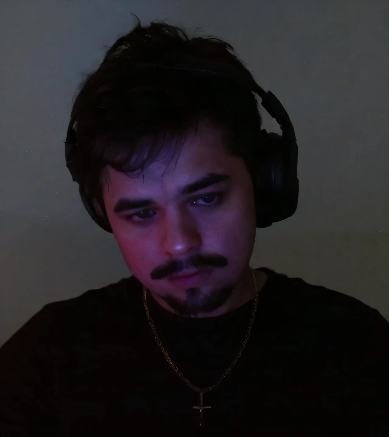 The image appears to show a person with short, dark hair, a mustache, and a goatee, wearing a dark shirt and a necklace with a cross pendant. The individual is wearing headphones and is in a dimly lit room with a soft, reddish-purple glow on their face. They have a calm, focused expression while looking slightly downward.