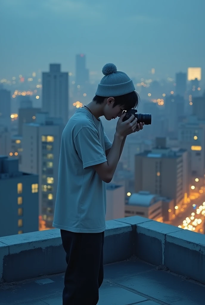 The image shows an adult Japanese male sitting perched on a rooftop at dusk or nighttime with a cityscape in the background. The individual is dressed casually, with a light grey beanie hat obscuring his face, a loose fitting white T-shirt, and black pants. His hair is short and obscured, and his face is tilted downwards, obscuring his expression. He is concentrating on adjusting the camera in his hands, seemingly preparing to take a picture. The scene has a cool, muted colour scheme, with the lighting casting a blue hue over the rooftop area, while the city below is lit with a warm yellow light from the buildings and cars in the parking lot. The overall atmosphere conveys a sense of quiet concentration and urban exploration.