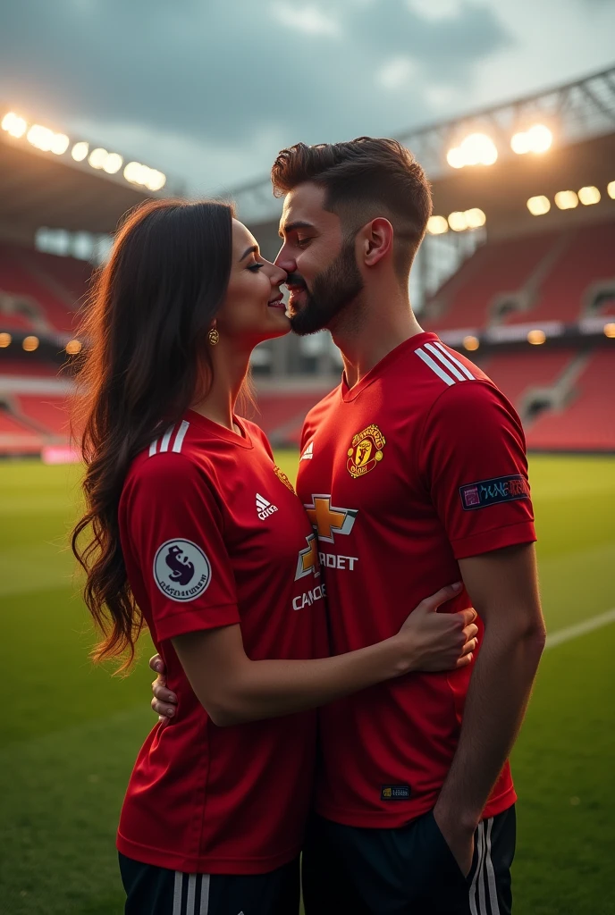 A man and a woman in Manchester shirts are on a football pitch when he kisses her on the cheek.