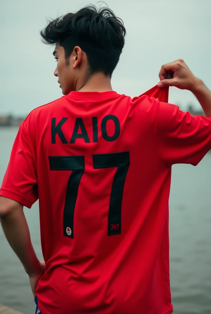 A young man with his back to the camera wearing a red shirt that says KAIO with the number 77. He is holding the shirt in his hand and showing the front of the shirt.