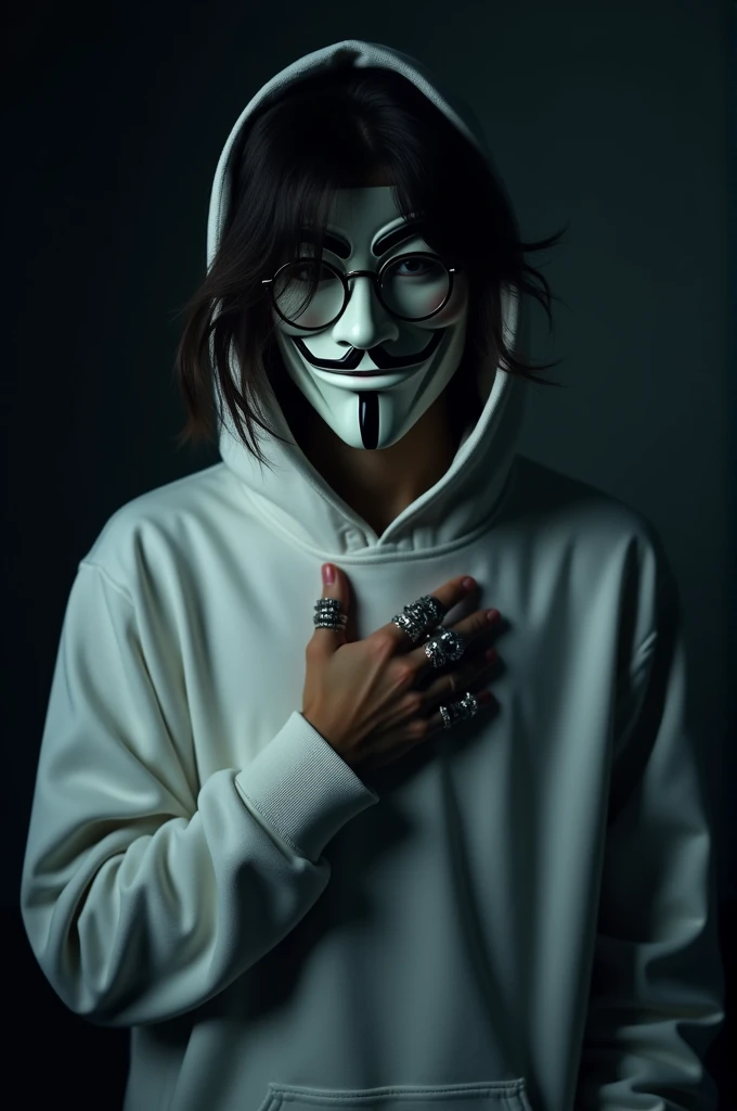A person with medium-length, messy dark hair is wearing a white hoodie and a stylized Guy Fawkes mask. The mask covers their entire face, and they are also wearing round eyeglasses over the mask. The individual is holding their hand up to their chest, with several rings visible on their fingers. The lighting in the image is dim, with a dark and moody ambiance, giving the entire scene a somewhat mysterious and rebellious atmosphere. The background is blurry and out of focus, emphasizing the person as the main subject of the image.